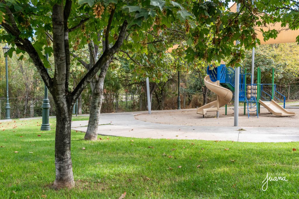 Plaza de juegos infantiles en San Carlos de Apoquindo, con resbalines, árboles frondosos y áreas verdes, ideal para niños y familias.